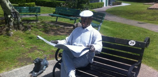 A man in a white buisness shirt and white flat cap sits on a black bench in a park and reads a newspaper