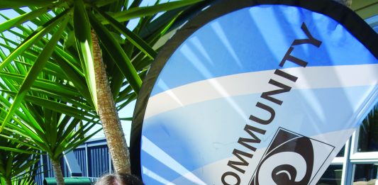 Picture of a young woman in a blue dress holing a peice od A3 card in front of her next to a flag with Merivale Community Centre written on it.