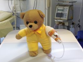 Picture of hospital teddy bear in yellow shirt with a drip on its arm sitting on a hospital bed