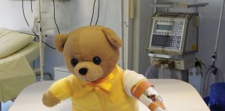 Picture of hospital teddy bear in yellow shirt with a drip on its arm sitting on a hospital bed