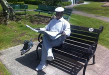 A man in a white buisness shirt and white flat cap sits on a black bench in a park and reads a newspaper